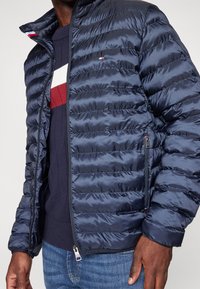 Granatowa kurtka puchowa zapinana na suwak, z poziomym pikowaniem, dzianinowymi mankietami i subtelnymi akcentami z logo. Noszona na granatowym i czerwonym swetrze.