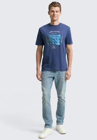 Granatowa koszulka z krótkim rękawem z graficznym wzorem, na którym znajduje się niebieski kwadrat i tekst. Połączona z jasnoniebieskimi dżinsami i białymi trampkami.