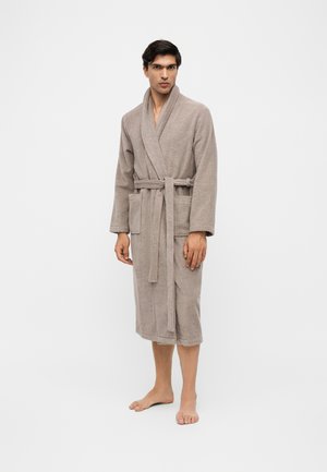 Homme debout pieds nus portant un long peignoir beige noué à la taille avec deux poches avant sur un fond blanc uni.