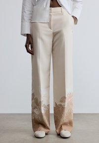 Bēšas bikses ar ziedu rakstiem rozā un brūnā krāsā apakšā. Izgatavotas no gludas auduma, ar piegriezumu pēc pasūtījuma un platām biksēm.