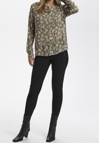 Olijfgroene blouse met lange mouwen en knopen, met een wit bloemenpatroon, gecombineerd met zwarte skinny broek en zwarte enkelhoge laarzen met hak.