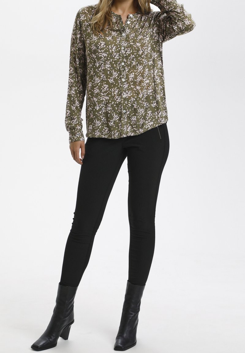 Olijfgroene blouse met lange mouwen en knopen, met een wit bloemenpatroon, gecombineerd met zwarte skinny broek en zwarte enkelhoge laarzen met hak.