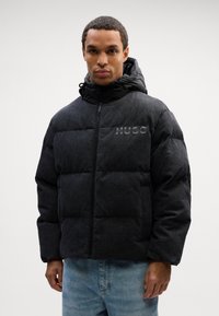 Svart pufferjakke med blankt hardware, quiltet design og høy hals. Har hette og en subtil logo på brystet. Kombinert med blå jeans.