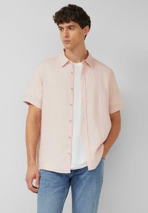 Junger Mann trägt ein hellrosa kurzärmeliges Hemd über einem weißen T-Shirt und blauen Jeans, schaut zur Seite mit einer Hand in der Tasche.