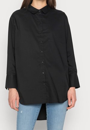 Femme portant une chemise noire ample à manches longues boutonnée, associée à un jean bleu clair déchiré, se tenant devant un fond uni.