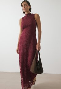 Burgundyröd spetsklänning med hög hals och ärmlös design; har intrikata blommönster och en sidoklyvning; kompletteras av en brun handväska.