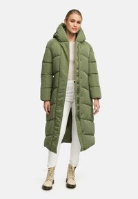Piumino verde con cappuccio lungo e oversize, design trapuntato, bottoni frontali e taglio dritto, abbinato a pantaloni chiari e stivali.
