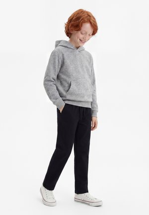 Enfant roux souriant, portant un sweat à capuche gris, un pantalon noir et des baskets blanches, debout avec une main dans la poche sur fond blanc.