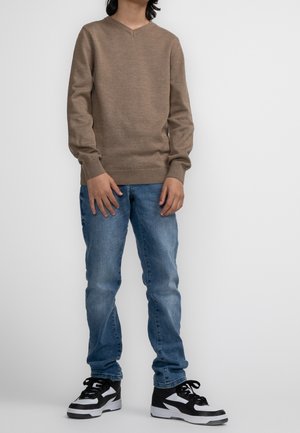 Pull en maille marron avec un col en V, associé à un jean bleu clair et des baskets noires et blanches. Le pull présente des poignets et un ourlet à côtes.