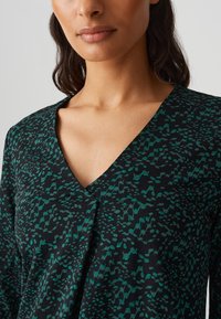 Top nero a scollo a V con una texture astratta in tonalità turchese. Tessuto morbido con una vestibilità rilassata, che mette in risalto la scollatura e i dettagli del design.