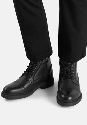 Czarne skórzane botki, które mają teksturowane wykończenie, sznurowany fason, gumową podeszwę oraz zamek błyskawiczny po bokach. Doskonale pasują do czarnych spodni.