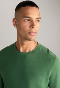 Groen textuur gebreide trui met een ronde hals. Close-up weergave toont het gedetailleerde stofpatroon en de zachte, ademende textuur.