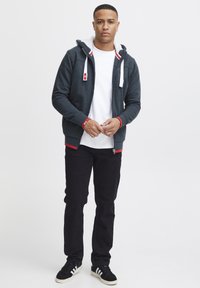 Dunkelgrauer Reißverschluss-Hoodie mit weißem Kunstfellfutter, roten Akzenten, kombiniert mit einem weißen T-Shirt und schwarzen Hosen, dazu schwarze Sneaker.