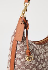 Braune Handtasche mit strukturiertem Körper, Lederakzenten, goldfarbener Hardware und einem markanten Logo-Tag, das an dem Tragegurt befestigt ist.