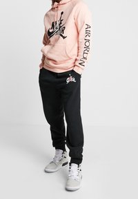 Světle růžová mikina s černým grafickým nápisem "Nike Air"; černé tepláky s malým logem. Šedé tenisky s texturovaným vzorem a bílými tkaničkami.