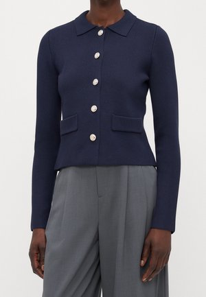 Femme portant un cardigan bleu foncé boutonné avec des boutons argentés et deux poches à rabat à l'avant, associé à un pantalon large gris taille haute.