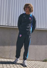 Black athletic tracksuit with blue accents, featuring team emblem. Texture is smooth, pants have elastic cuffs, worn with gray sneakers.