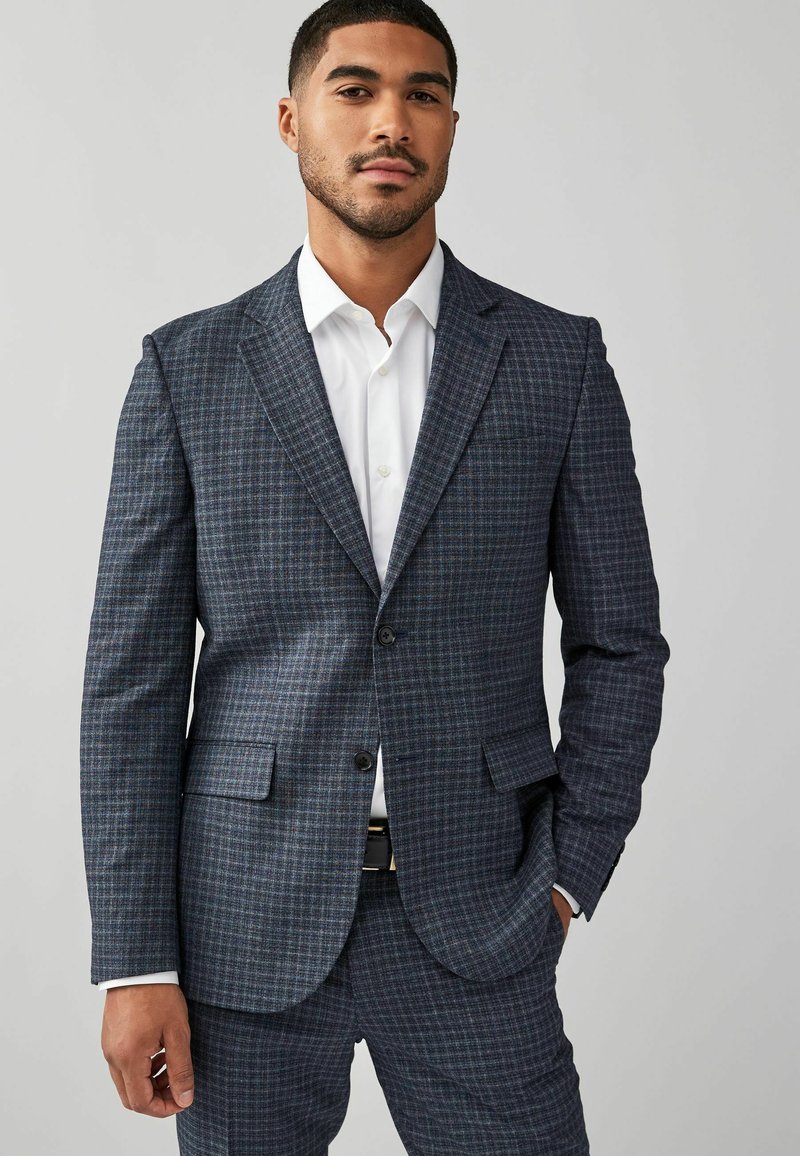 Marineblauw geruite blazer van gestructureerde stof, met gelapte revers, twee knopen en twee voorzakken; gedragen over een wit shirt.