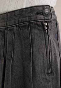 Szürke farmernadrág redőkkel, cipzáras záródással és kerek logó gombbal. Jellemzője a textúrált felület és a strukturált derékpánt.