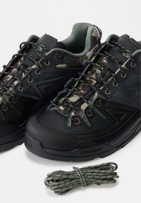 Μαύρα και καμουφλαρισμένα ορειβατικά παπούτσια με δεμένα κορδόνια, επενδεδυμένος γιακάς και επιπλέον πράσινα κορδόνια τυλιγμένα δίπλα το υς, σε λευκό φόντο.