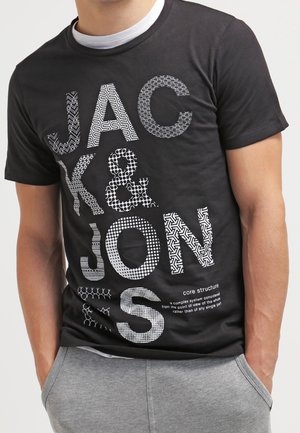 Homme portant un t-shirt noir avec "JACK & JONES" en grandes lettres blanches à motifs et un pantalon de survêtement gris, les mains dans les poches.