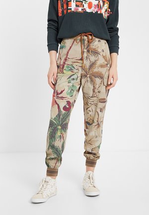 Personne portant un pantalon de jogging beige avec un imprimé palmiers tropicaux, taille avec cordon marron, poignets élastiques, baskets blanches montantes et sweat-shirt foncé.