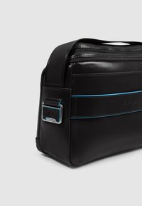 Zwarte leren boodschappentas met blauwe accentstiksels, een ritssluiting aan de bovenkant en een zilverkleurig hardwaredetail aan de zijkant. Soepele textuur en rechthoekige vorm.