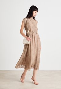 Rjava midi obleka v ribje kost prileganjem v pasu, s kapicami na rokavih in ohlapnim krilom, združena z majhno belo torbico in golimi sandali na peto.