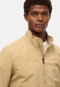 Beige jakke med ståkrave, frontlynlås og glat tekstur. Designet med en strømlinet pasform og minimale sømdetaljer.