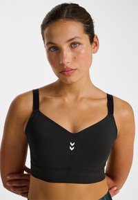 Schwarzer Sport-BH aus weichem Stoff, mit V-Ausschnitt, verstellbaren Trägern und einem weißen Chevron-Logo in der Mitte.