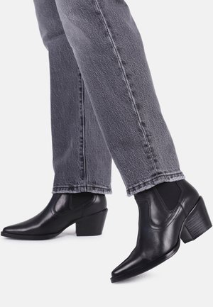 Personne portant des bottines en cuir noir à talons blocs et un jean droit gris foncé sur un fond uni.