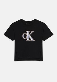 Camiseta corta negra de algodón, con un gran logo "ck" en gris y blanco con acentos metálicos. Mangas cortas, con aberturas laterales.