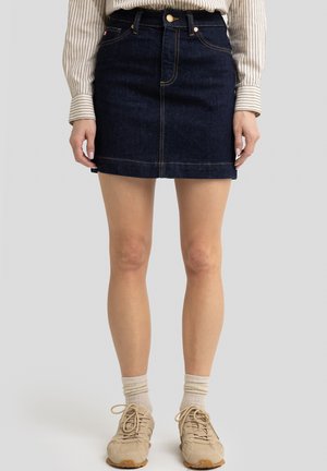 Dunkelblaue Denim-Minirock mit gelben Nähten, vorderen Taschen und klassischer Passform. Kombiniert mit beigen Sneakern und hellen Socken.