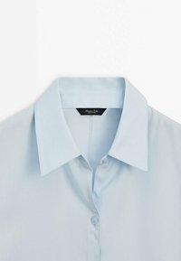 Chemise bleu clair à col avec boutons, soigneusement pliée et présentée sur un fond blanc.