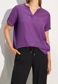 Top viola a maniche corte con scollatura a V e risvolti, realizzato in tessuto testurizzato e dalla vestibilità ampia, indossato con pantaloni neri con stringa in vita.