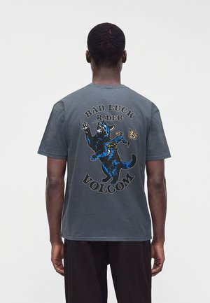 Homme de dos portant un t-shirt gris foncé avec un chat de dessin animé coloré et le texte "Bad Luck Rider Volcom" dans le dos.