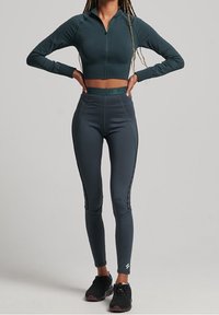 Chaqueta corta con cremallera de color verde oscuro y leggings de cintura alta gris oscuro con acentos laterales, combinados con zapatillas deportivas negras.