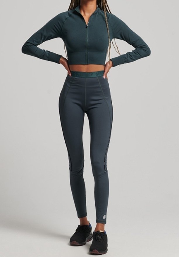 Chaqueta corta con cremallera de color verde oscuro y leggings de cintura alta gris oscuro con acentos laterales, combinados con zapatillas deportivas negras.