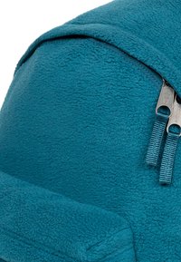 Mochila de felpa en color verde azulado con dos tiradores de cremallera, forma redondeada y textura suave. Incluye un panel trasero acolchado para mayor comodidad.