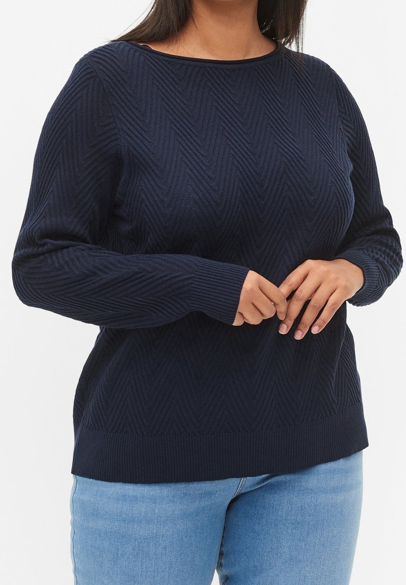 Mörkblå stickad tröja med ett strukturerat chevronmönster, långa ärmar och rund halsringning, i kombination med ljusblå denimbyxor.