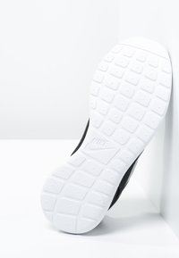 Semelle de sneaker Nike blanche avec un motif de semelle carré et le logo Nike, inclinée contre des murs et un sol blancs.