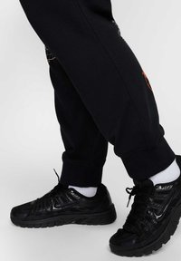 Pantalones de chándal negros con un ajuste ceñido, puños elásticos y un gráfico impreso. Llevados con zapatillas deportivas negras y calcetines blancos hasta el tobillo.