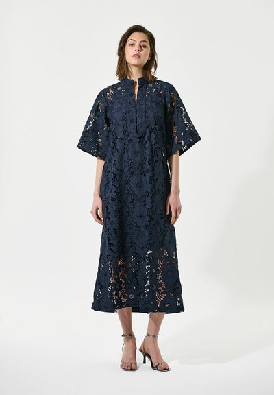 Femme portant une robe midi en dentelle florale bleu marine à manches courtes, debout en sandales à talons hauts sur un fond uni.