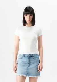 Femeie cu părul drept și închis la culoare, purtând un tricou alb ajustat și o fustă din denim de culoare albastră deschisă, cu tiv destrămat, stând în fața unui fundal alb.
