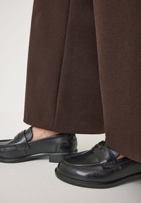 Schwarze Leder-Lederschuhe mit glatter Oberfläche, ausgestattet mit einem dekorativen Riemen und minimalen Akzenten, kombiniert mit weiten braunen Hosenbeinen.