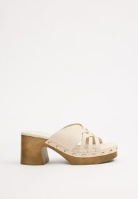 Sandalias de plataforma beige con tacón de bloque de madera, que presentan tiras entrecruzadas y acentos de remaches en tono dorado a lo largo del borde de la suela.