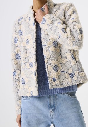 Personne portant un jean bleu clair et une veste blanche texturée avec de grands motifs floraux bleus et beiges, par-dessus un pull bleu.