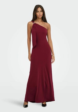 Femme portant une robe bordeaux sans manches, longue jusqu'au sol, avec une bretelle et des talons noirs sur un fond clair.