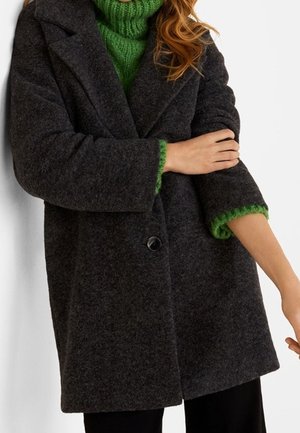 Vrouw die een donkergrijze wollen jas draagt over een felgroene, dik gebreide coltrui, met haar armen over haar borst gevouwen.