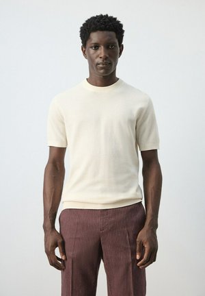 Mand stående iført en cremefarvet kortærmet sweater og brune bukser med smalle striber mod en ensartet lysegrå baggrund.